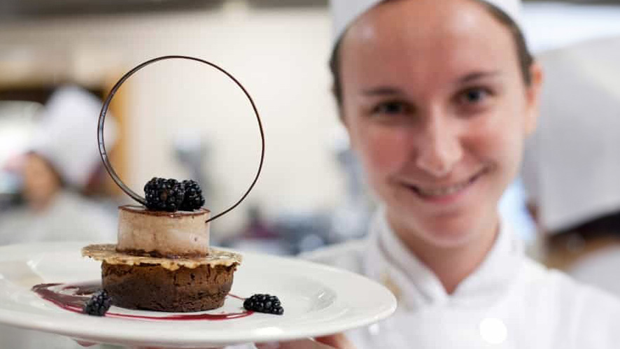 A CIA student presents a dessert that includes a chocolate base, a light-colored topping, blackberries, and a decorative chocolate ring.