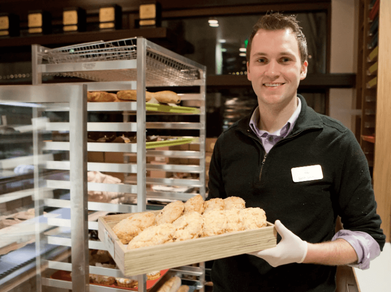 A smiling person holds a tray of pastries in a bakery, showcasing the delicious results of pastry vs baking on racks behind them.