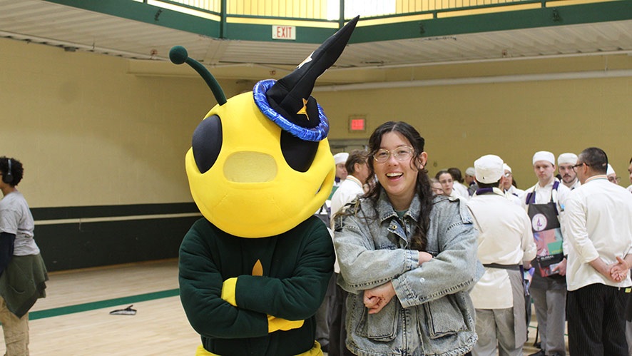 Image of Sting, CIA’s bee mascot and a smiling CIA student stand with arms crossed in the gym during the fifth annual Tri-Wizard Tournament at CIA in Hyde Park, NY.