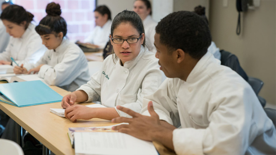 CIA students in chef uniforms talking and reading in a classroom.