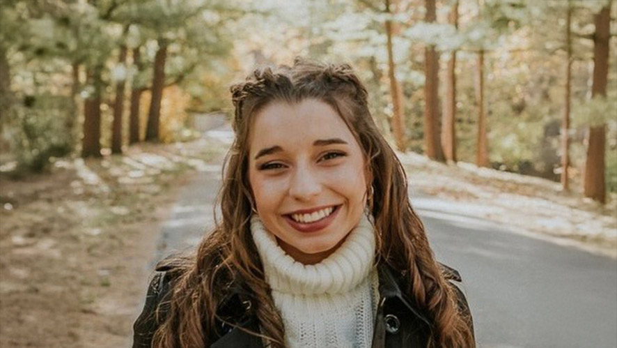 CIA student Ava Swank in a sweater and jacket standing on a wooded road outdoors.